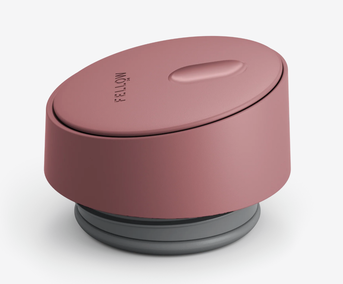 En pink, oval Fellow Carter Slide Mug termo krus med "FELLOW" på toppen, en tekstureret knap og en grå cirkulær base vises på en hvid baggrund.