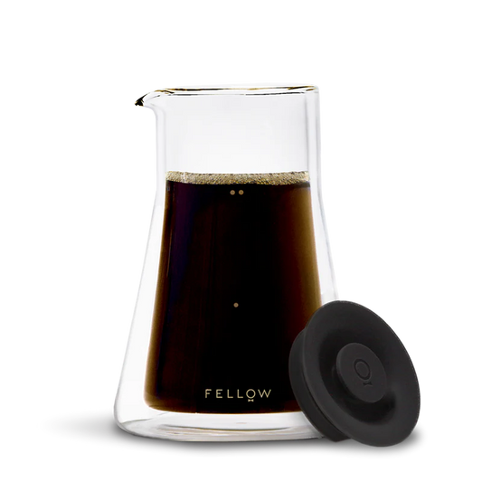 En gennemsigtig Fellow Termo Karaffel af glas fyldt med mørk kaffe har en tud på toppen og "Fellow"-etiketten i bunden. Et rundt sort låg komplementerer dets slanke design mod en almindelig sort baggrund.