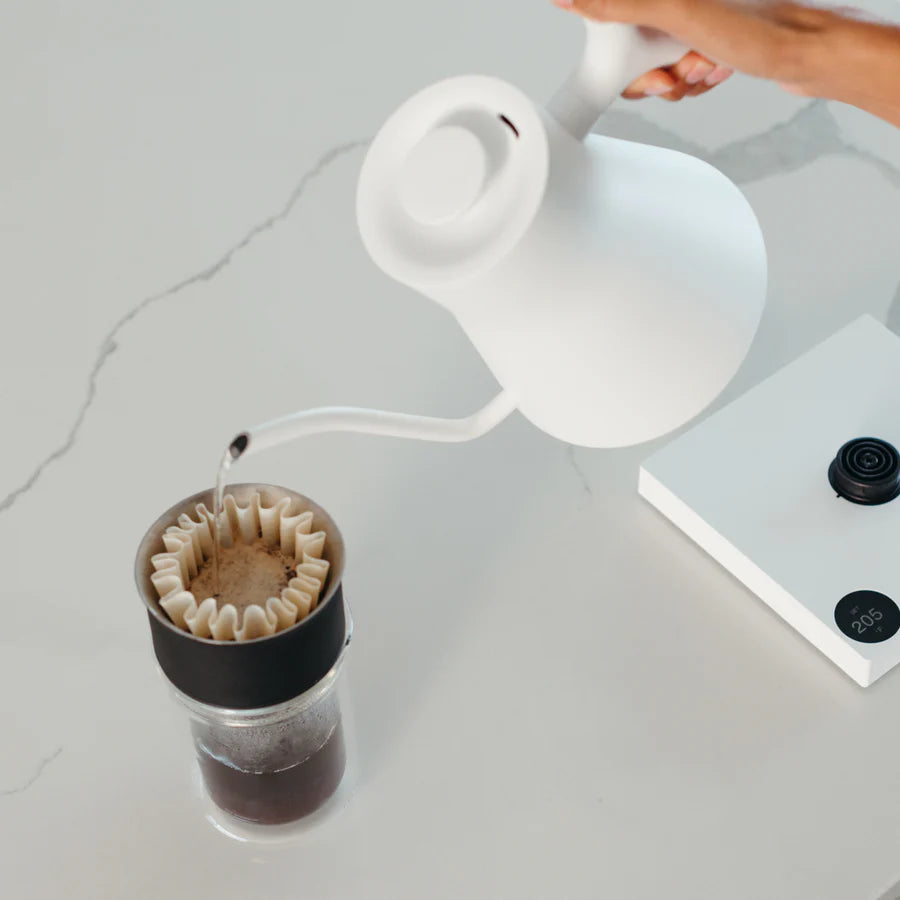 En hånd hælder vand fra en Fellow Stagg EKG PRO 0,9 L elkedel i en kaffedrypper med et papirfilter over en glasbeholder med en digital vægt på en hvid bordplade.