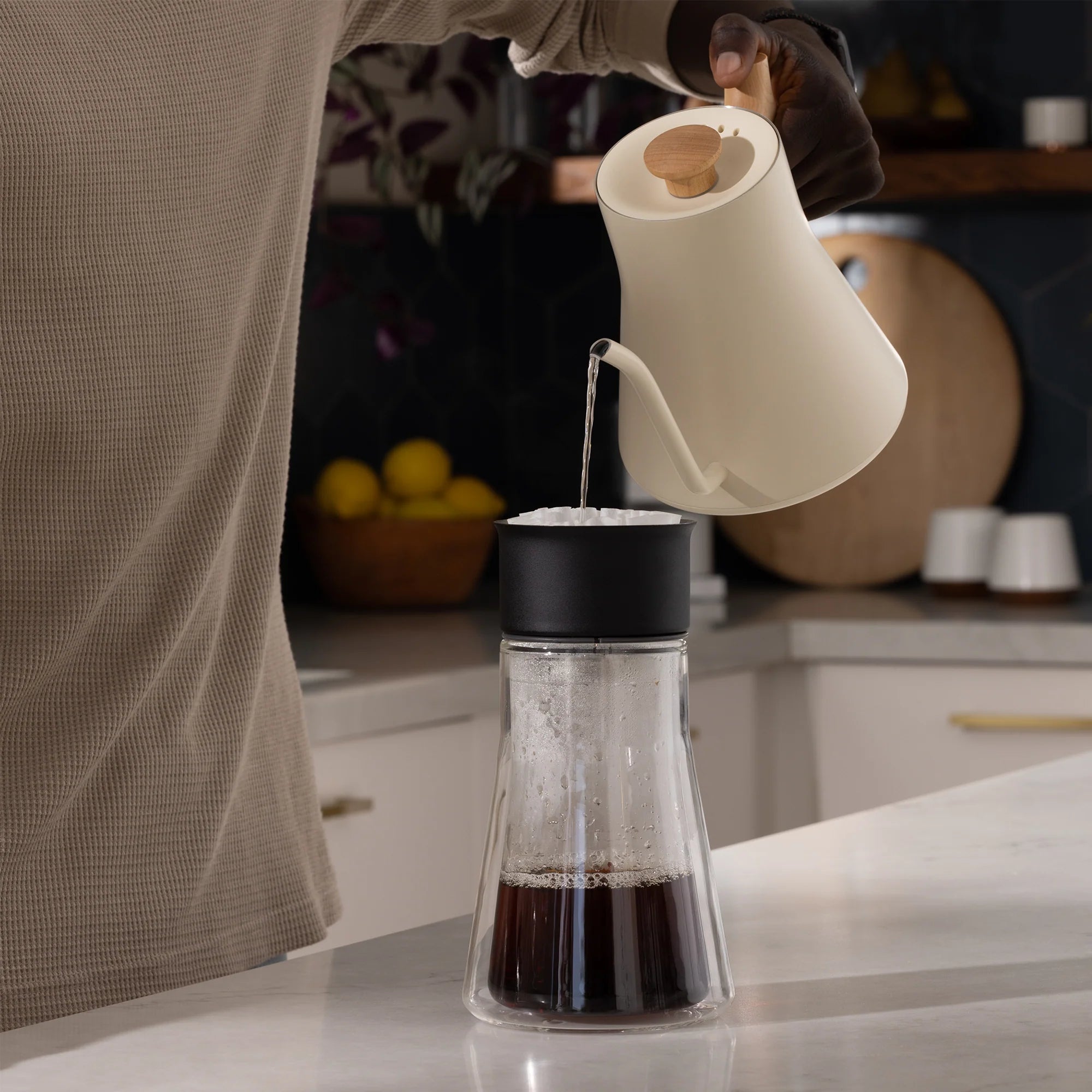 En person hælder varmt vand fra en Fellow, Stagg EKG 0,9 L elkedel med en præcisions-tud i en sort og glas kaffedryp på disken, omgivet af citroner og fade.