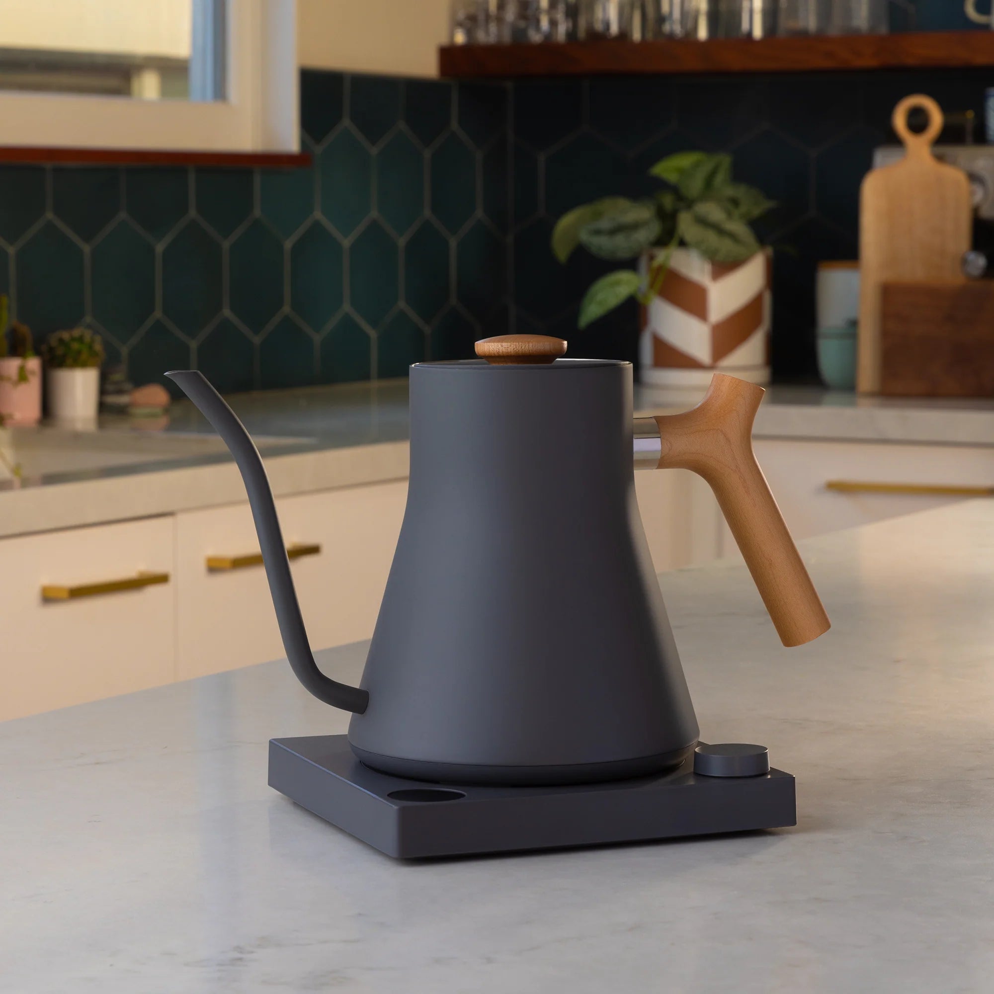 Fellow Stagg EKG 0,9 L Elkedel fra Fellow har med sit slanke sorte design og træhåndtag en præcisions-tud og temperaturkontrol. Den er sat på en køkkenbordplade med en bagplade med grønne fliser og forskellige køkkenartikler i baggrunden for perfekt brygning.