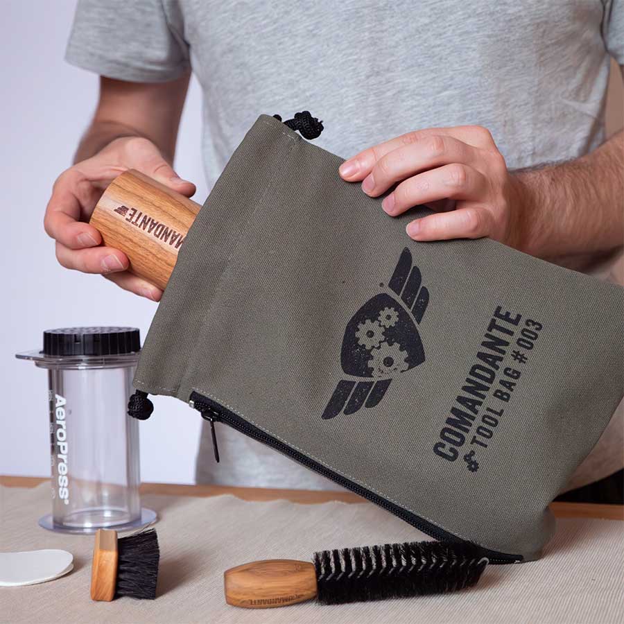 Nogen lægger en Comandante-kværn med træhåndtag ned i en grøn Comandante Toolbag #3. På bordet i nærheden står en AeroPress, en lille børste og en stor børste.