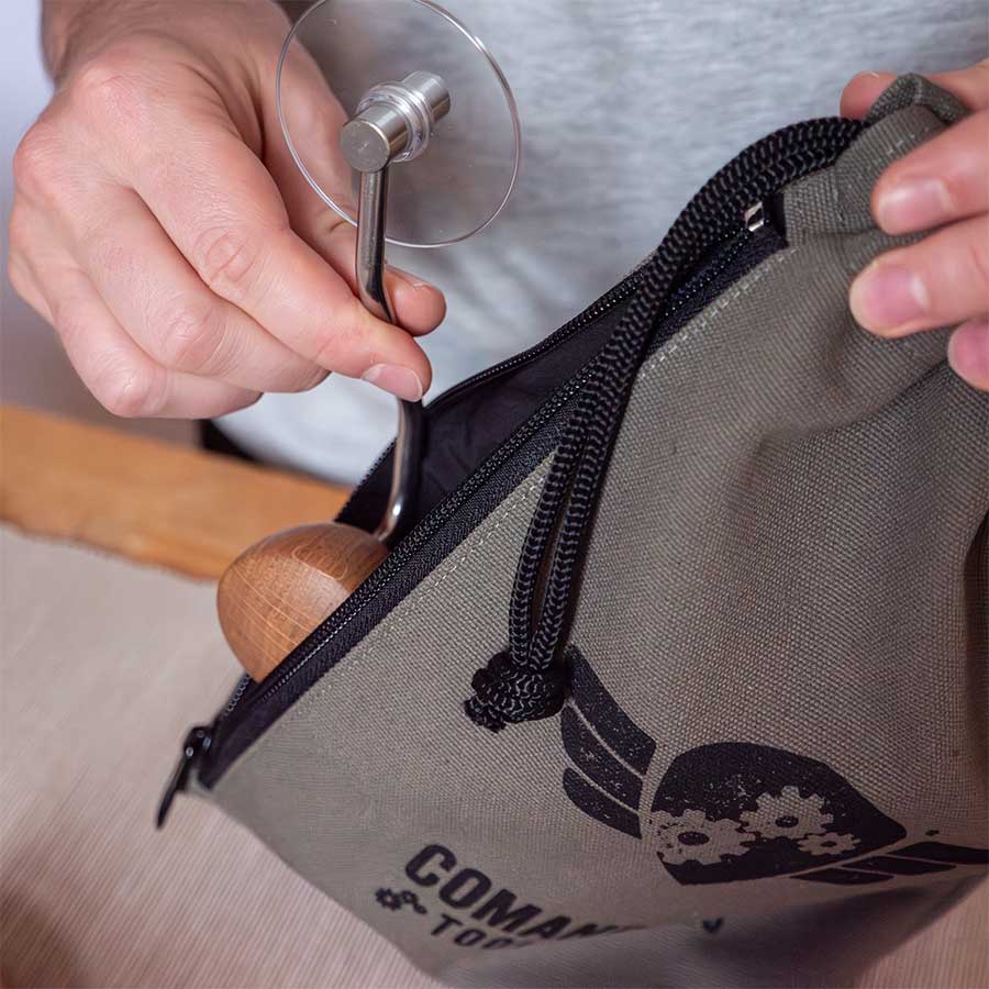 Nogen lægger en Comandante-kværn med træhåndtag og metalhåndsving i den grå Comandante Toolbag #3, som har geargrafik og "COMANDANTE"-logo.