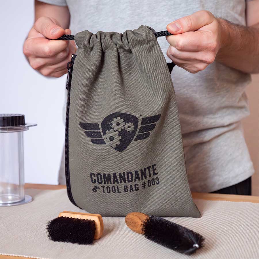 En person placerer en Comandante Toolbag #3 fra Comandante på et bord sammen med en kaffetaske, en lille pensel, en stor pensel og en klar cylindrisk beholder.