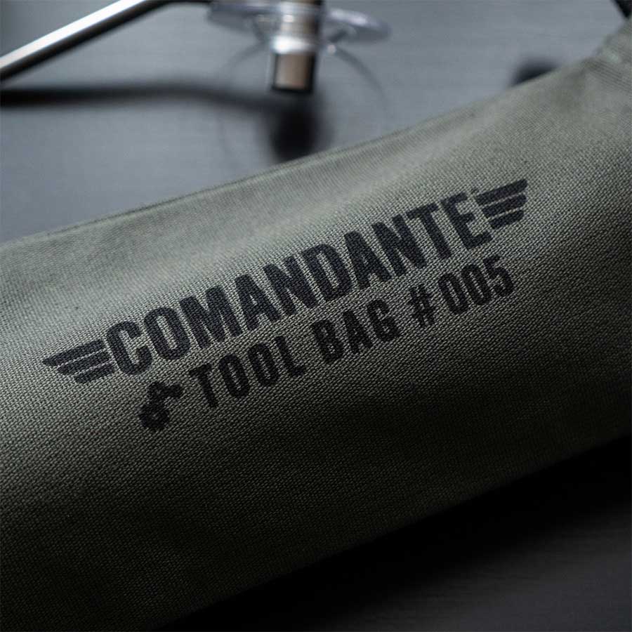 Et nærbillede af den grønne Comandante Toolbag #5 fra Comandante med "#005" og vingegrafik. Opbevaringsgenstande til kaffegrej i metal og plast ses i baggrunden.