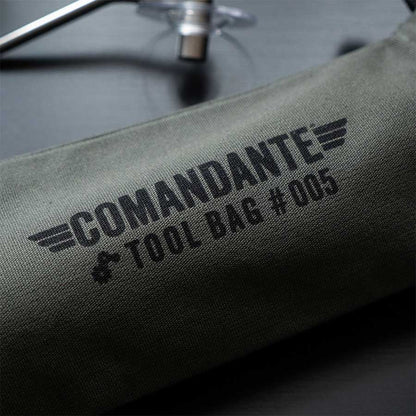 Et nærbillede af den grønne Comandante Toolbag #5 fra Comandante med "#005" og vingegrafik. Opbevaringsgenstande til kaffegrej i metal og plast ses i baggrunden.