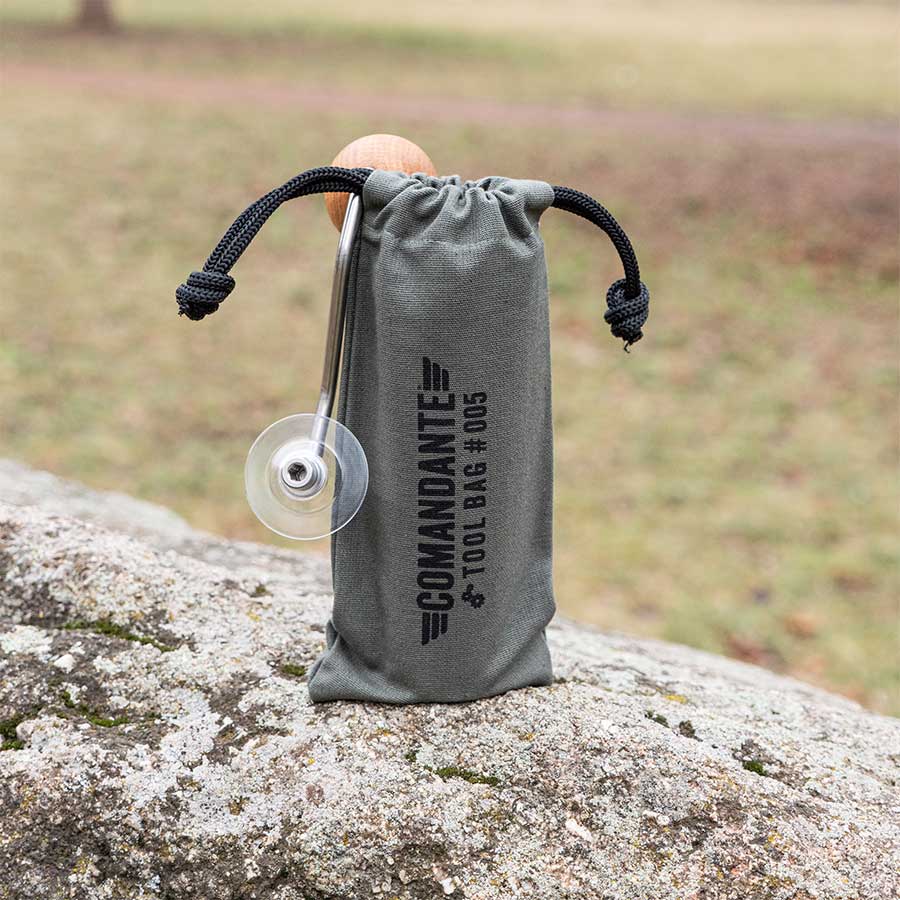 Den grå Comandante Toolbag #5 fra Comandante hviler på en sten udendørs og holder et metalredskab med en cirkulær klinge - ideel til opbevaring af dit kaffeudstyr. Den græsklædte baggrund er blødt sløret.