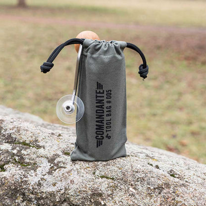 Den grå Comandante Toolbag #5 fra Comandante hviler på en sten udendørs og holder et metalredskab med en cirkulær klinge - ideel til opbevaring af dit kaffeudstyr. Den græsklædte baggrund er blødt sløret.