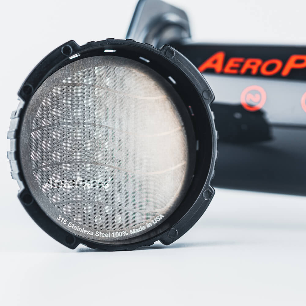 Nærbillede af et AeroPress Metalfilter med "316 Stainless Steel 100% Made in USA" på kanten sikrer holdbarhed og lang levetid. En del af AeroPress-kaffemaskinen med røde bogstaver er synlige i baggrunden, hvilket understreger dens genanvendelige design.