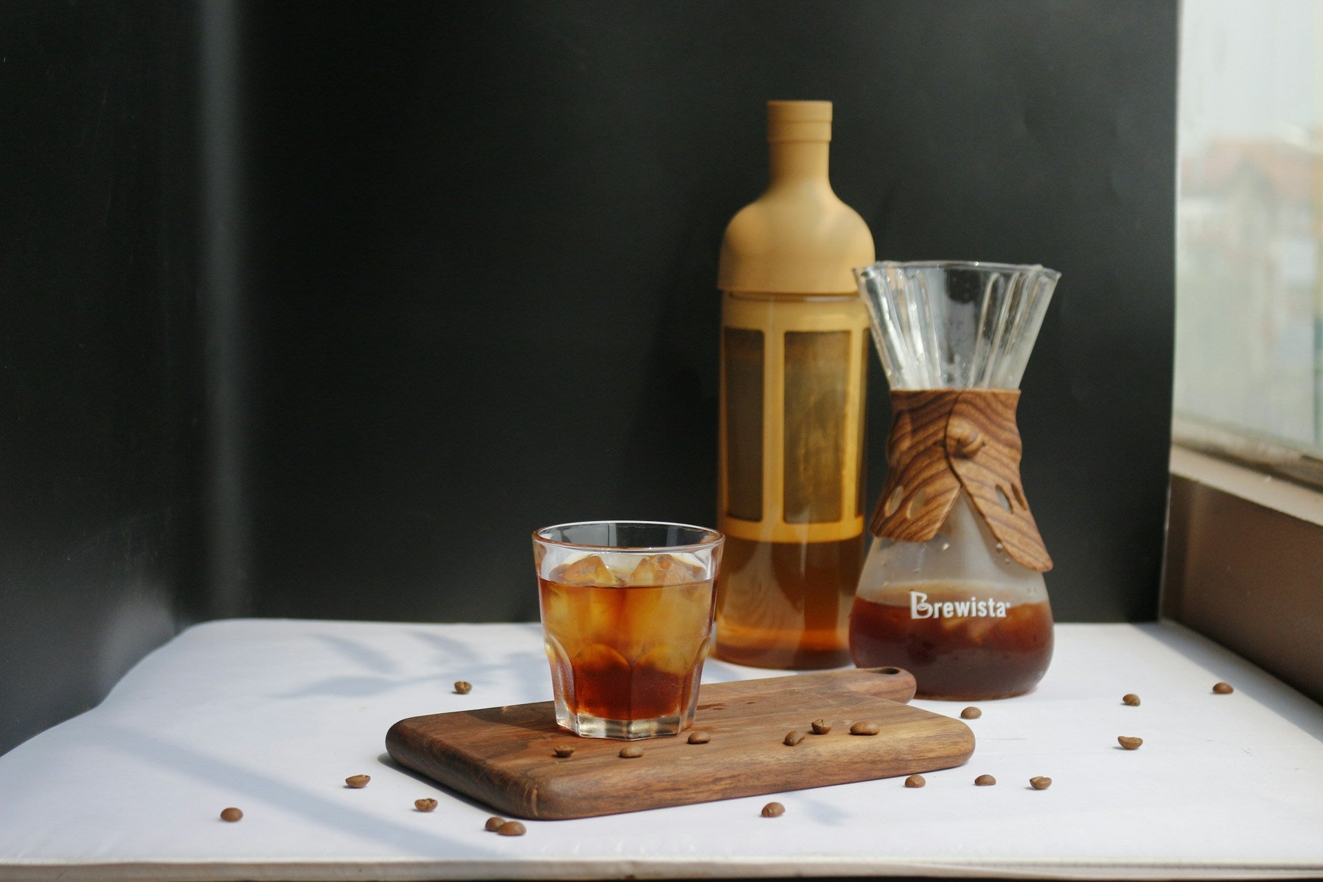 Et glas EverBean Limited ICED Coffee Blend lavet af Arabica-bønner står på en træplade med kaffebønner ved siden af en Brewista-karaffel og en flaske, alt sammen mod en mørk baggrund nær et vindue.