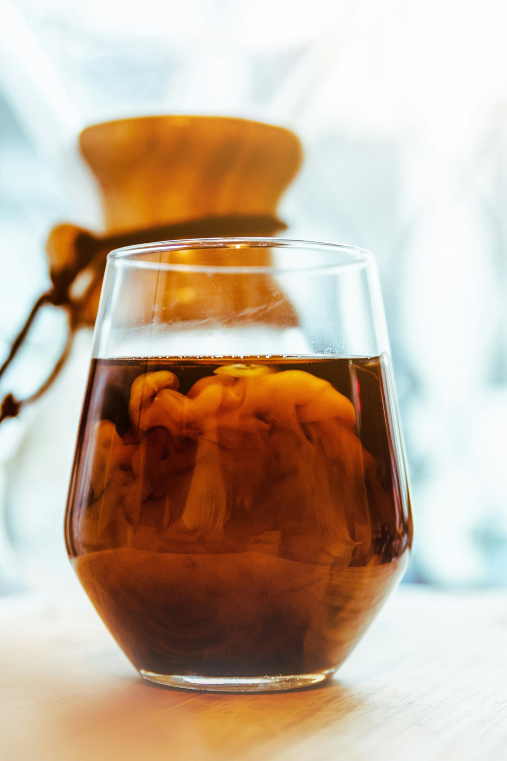 Et glas EverBean Limiteds ICED Coffee Blend, lavet med bløde Arabica-bønner og hvirvlende mælk indeni, hviler på en lys overflade med en sløret Chemex-kaffemaskine i baggrunden.