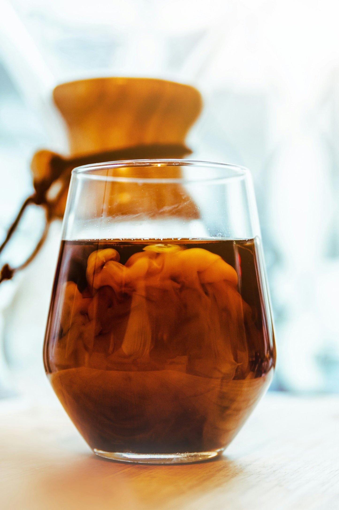 Et glas EverBeans ICED Coffee Blend (w) står på et træbord, med cremede hvirvler marmorerer det mørke Arabica-bønner-bryg. En Chemex kaffemaskine med sin trækrave sløres blødt i baggrunden.