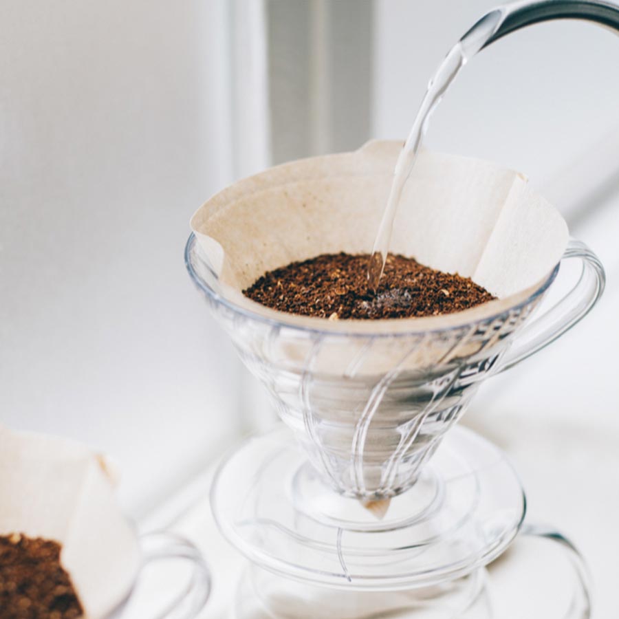 En pour-over-kaffemaskine i klart glas med en Hario Papirfiltre fyldt med malet kaffe i en 02-drypper, mens varmt vand hældes over for at starte brygningen.