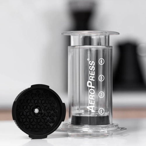 En Aeropress AeroPress-kaffemaskine med nummererede markeringer står oprejst ved siden af et AeroPress Flow Control Filter Cap-tilbehør fra Aeropress, der er ideelt til at opnå espressolignende crema. Baggrunden er blødt sløret.