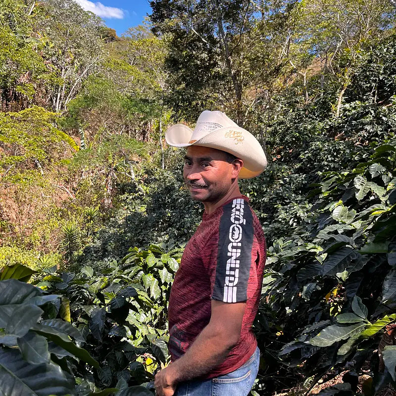 En mand med cowboyhat og rød skjorte står på en frodig, grøn eng, omgivet af planter og træer, og smiler til kameraet under en blå himmel.