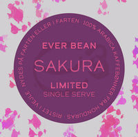 På en rund etiket med pink og lilla stænk står der: "EverBean Limited Single Serve, Sakura". Nyd praktisk single-serve kaffe lavet af 100% Arabica sakura-bønner fra Honduras, ristet i Viborg - perfekt til kaffe på farten.