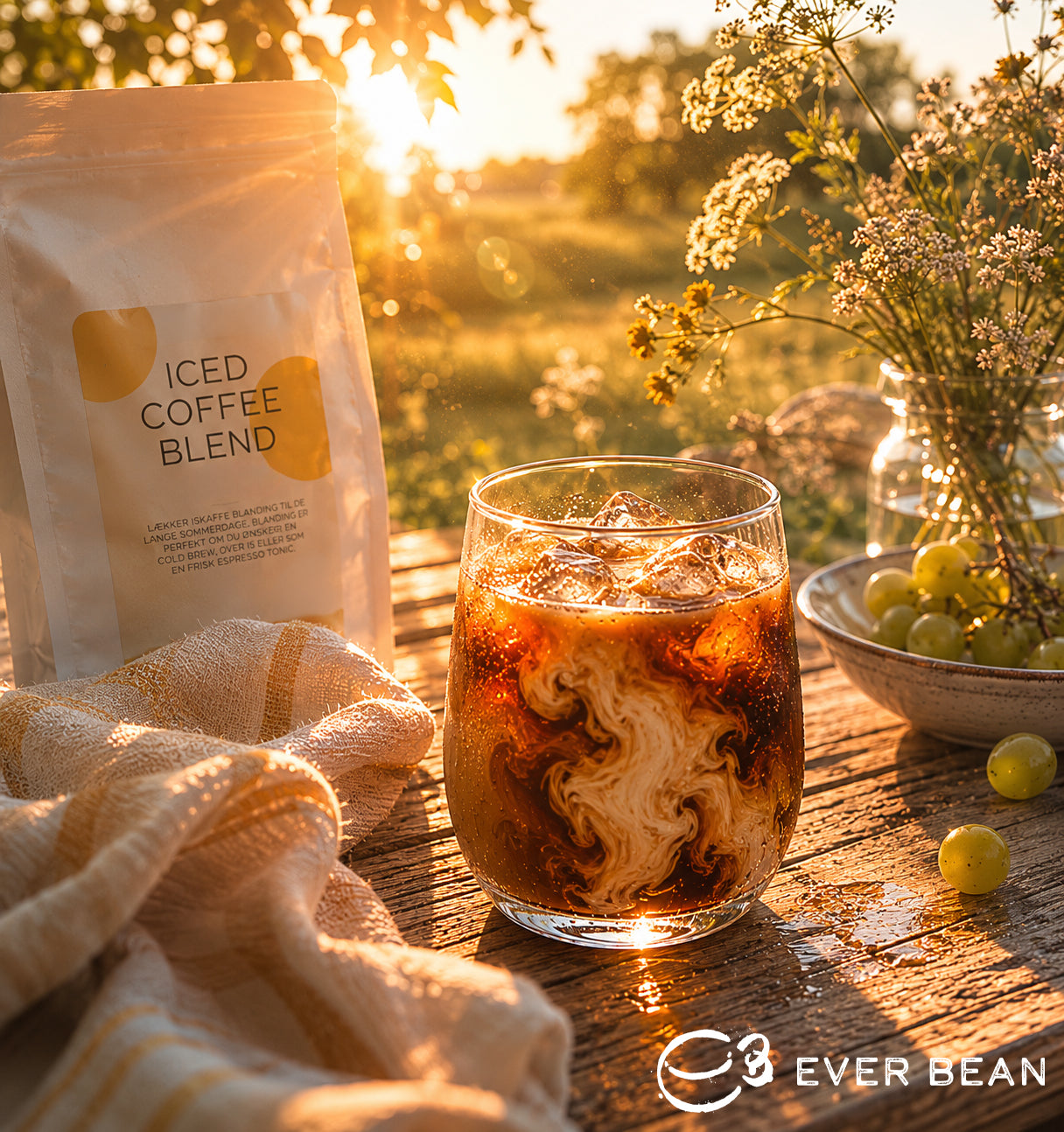 Et glas ICED Coffee med flødehvirvler står på et rustikt bord ved siden af en "ICED Coffee Blend"-pakke fra EverBean Limited, lavet af arabica-bønner, mens sollyset strømmer gennem en naturskøn udendørs baggrund.
